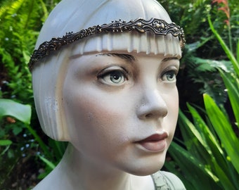 Könnte beinhalten: Ein silbernes Stirnband mit einem dekorativen Design, getragen von einer Puppe mit kurzen, glatten Haaren. Das Stirnband ist aus Metall und hat ein zartes, filigranes Muster.
