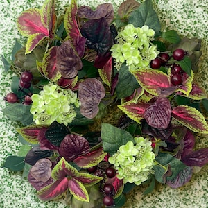 May include: A vibrant wreath featuring a variety of colorful foliage. The wreath includes green, purple, and red leaves, along with clusters of small, round, dark red berries and light green hydrangea flowers. The wreath is set against a speckled green background.