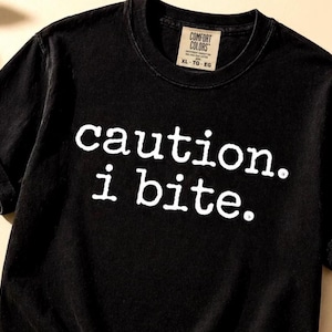 Puede incluir: Camiseta negra con el texto blanco "caution. i bite." en negrita. La camiseta tiene cuello redondo y mangas cortas. Una etiqueta en el cuello dice "COMFORT COLORS" y "XL - TG - EG".