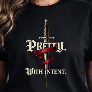 May include: Black t-shirt featuring a sword design with a red ribbon and the words "Pretty With Intent." The sword is gold-colored with a detailed hilt. The text is in a stylized font.
