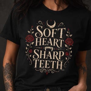 Könnte beinhalten: Schwarzes T-Shirt mit dem Text "SOFT HEART SHARP TEETH" in gotischer Schrift, umgeben von roten Rosen und Ranken. Über dem Text befindet sich ein Halbmond. Das Shirt hat kurze Ärmel.