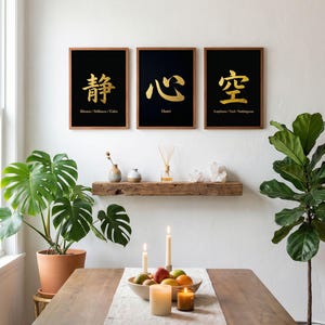 Op de afbeelding: Drie ingelijste kunstdrukken met zwarte achtergronden en gouden kalligrafie hangen boven een houten tafel. De prints tonen Aziatische karakters met Engelse vertalingen: Silence/Stillness/Calm, Heart en Emptiness/Void/Nothingness. Op de tafel staan kaarsen en een fruitschaal.