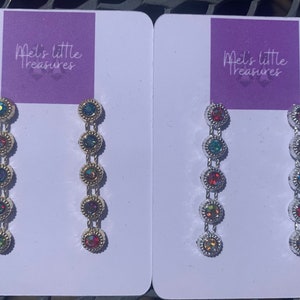 Pode incluir: Dois pares de brincos pendurados com pedras preciosas iridescentes e coloridas. Cada brinco tem cinco pedras redondas com moldura conectadas por correntes delicadas. Os brincos são exibidos em cartões brancos com o texto "Mel's little treasures".