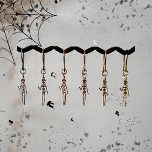 Könnte beinhalten: Sechs Skelett-Ohrhänger in Silber-, Kupfer- und Goldtönen. Jeder Ohrring hat einen detaillierten Skelettanhänger, der an einem einfachen Haken hängt. Die Ohrringe werden vor einem strukturierten Hintergrund mit Spinnennetz-Design präsentiert.