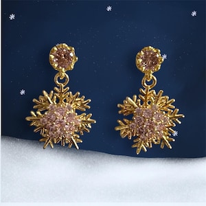 May include: A pair of gold-colored snowflake earrings with pink gemstones. Each earring features a round pink gem at the top, connected to a detailed snowflake design with more pink gems in the center. The background is a dark blue with white star accents.