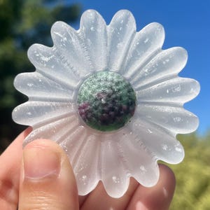 May include: A clear, daisy-shaped crystal with a green and red center. The flower has multiple petals and is held up against a blue sky. The crystal is a decorative item.