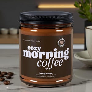 May include: A brown glass jar candle with a black lid. The label reads "Cozy Morning Coffee" with the scent notes of coffee, cream, and vanilla. The candle is soy and coconut wax, handmade in Lafayette, LA.