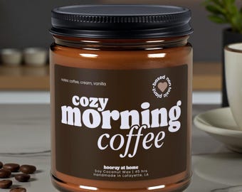 May include: A brown glass jar candle with a black lid. The label reads "Cozy Morning Coffee" with the scent notes of coffee, cream, and vanilla. The candle is soy and coconut wax, handmade in Lafayette, LA.
