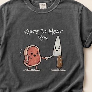 Peut inclure: T-shirt gris foncé avec l'inscription "Knife to Meat You" et une illustration de dessin animé. L'illustration montre un steak et un couteau se serrant la main. Le steak a une expression triste et le couteau a un visage souriant.