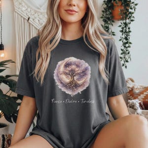 Può includere: T-shirt grigio scuro con un design celeste. Il design presenta un albero all'interno di una luna crescente, su uno sfondo viola e bianco. Il testo "Fierce. Divine. Timeless" è stampato sotto. La maglietta è in materiale morbido.