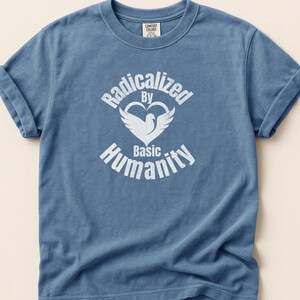 May include: Blue t-shirt with rolled-up sleeves. The shirt features the text "Radicalized By Basic Humanity" in white, with a dove and heart graphic. The shirt is made of a soft material.