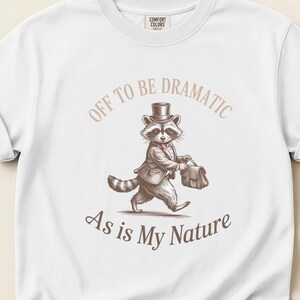 May include: White sweatshirt with a raccoon illustration wearing a suit and top hat, carrying a briefcase. The text reads "OFF TO BE DRAMATIC As is My Nature". The sweatshirt has a crew neck and the Comfort Colors logo.