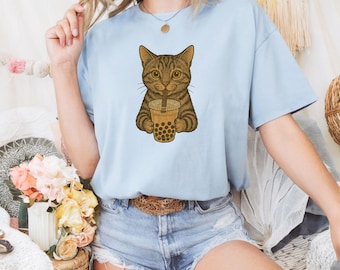 May include: Pale blue t-shirt with a brown tabby cat graphic drinking bubble tea. The cat is depicted with detailed fur and is holding a boba tea cup. The bubble tea cup is light brown with dark brown tapioca pearls.