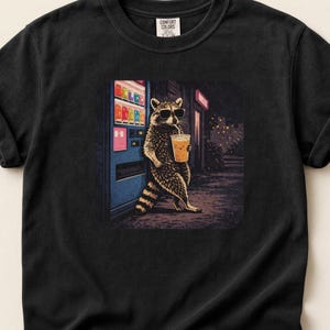 Może przedstawiać: Czarny t-shirt z grafiką szopa pracza w okularach przeciwsłonecznych, trzymającego napój ze słomką i opierającego się o automat. Szop pracz jest przedstawiony w szczegółowym, artystycznym stylu. Koszulka ma metkę Comfort Colors.