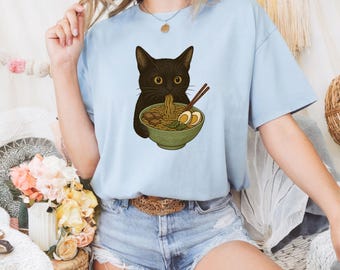 May include: Light blue t-shirt featuring a black cat eating ramen from a green bowl. The cat has yellow eyes and is holding chopsticks. The ramen bowl contains noodles, meat, and eggs. The shirt is a casual, oversized fit.