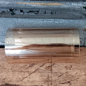 May include: A clear, cylindrical glass object with a curved shape, reflecting the surrounding environment. The glass is positioned on a weathered wooden surface, with a backdrop of distressed blue and orange painted wood.