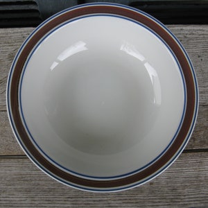 May include: A round ceramic bowl with a smooth white interior and a wide rim featuring a dark brown band bordered by two thin blue lines. The bowl has a simple, clean design with a glossy finish, showcasing a classic circular shape and neutral colors.