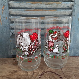 May include: Two clear glass drinking glasses with illustrations of the Twelve Days of Christmas. The glasses feature a cow and a maid, and a bird and a maid. The glasses have musical notes around the bottom.