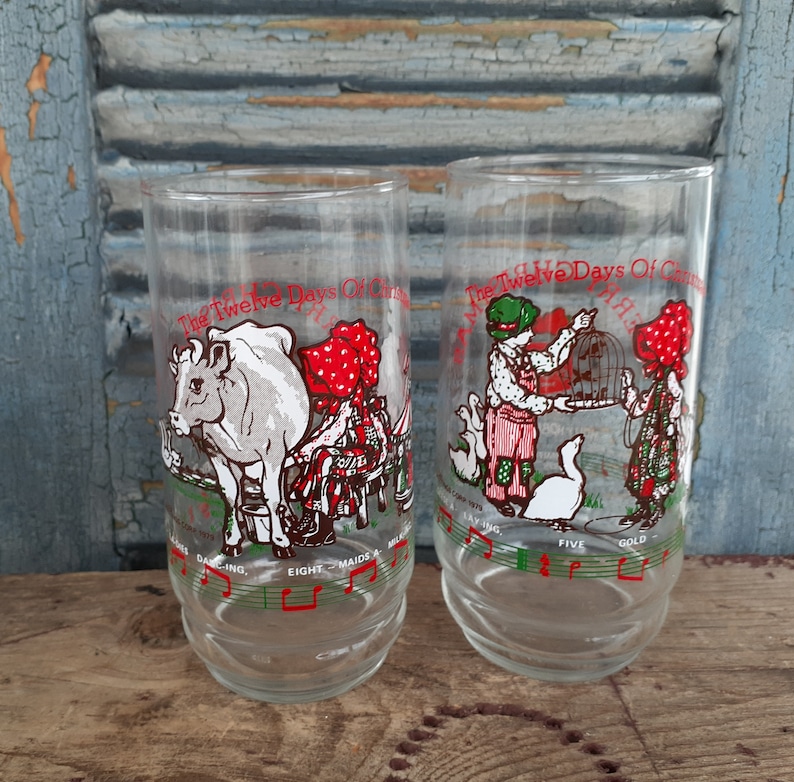 May include: Two clear glass drinking glasses with illustrations of the Twelve Days of Christmas. The glasses feature a cow and a maid, and a bird and a maid. The glasses have musical notes around the bottom.