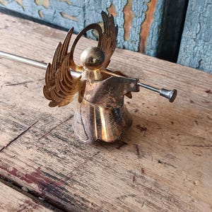 May include: A vintage brass angel figurine with outstretched wings and a trumpet. The angel has a detailed design and is set against a weathered wooden surface. The angel's wings are a warm gold color.
