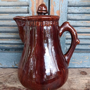 May include: A glossy, brown ceramic pitcher with a lid and handle. The pitcher has a spout for pouring and a decorative band near the top. The handle is arched, and the lid has a small knob. The pitcher is likely vintage or antique.
