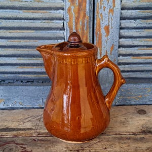 May include: A brown ceramic pitcher with a lid and handle. The pitcher has a glossy finish and a decorative band around the top. The handle is curved and has a textured design. The pitcher is likely used for serving beverages.