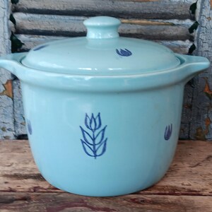 May include: A light blue ceramic casserole dish with a lid. The dish has two handles and is decorated with dark blue floral designs. The lid has a small knob. The casserole dish is on a wooden surface.