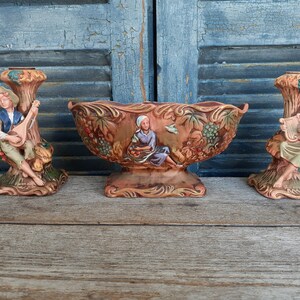 May include: A set of three ceramic decorative pieces featuring a central oval bowl and two matching candle holders. Each piece is finished in a warm brown glaze with hand-painted figures, grape clusters, and foliage motifs in blue, green, and tan.