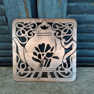 Puede incluir: Un posavasos de tetera de metal de color cobre con un diseño recortado de una tetera y una flor. La tetera tiene una tapa y un pico. La flor tiene cinco pétalos.