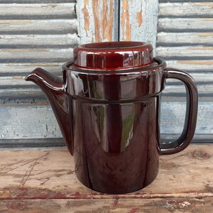 May include: A dark brown ceramic teapot with a matching lid and handle. The teapot has a spout for pouring and a cylindrical body. The glossy finish reflects light, highlighting its shape. The teapot is likely vintage or antique.