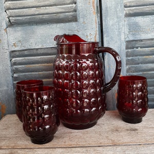 May include: A vintage red glass drinkware set featuring a pitcher and four matching glasses. The pitcher has a handle and a textured, bubble-like pattern. The glasses are also textured and have a similar design. The set is displayed on a wooden surface.