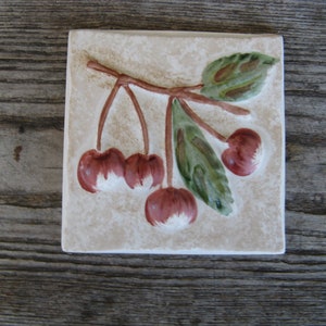 May include: A ceramic tile with a raised design of a cherry branch with four red cherries and green leaves.
