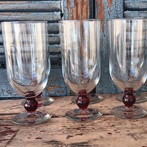 May include: Three clear glass goblets with a rounded base and a dark red ball-shaped stem. The goblets are displayed on a wooden surface against a weathered blue background.
