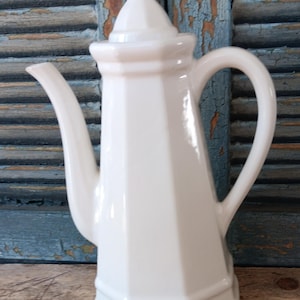 May include: A white ceramic coffee pot with a lid and a handle. The pot has a geometric, faceted design and a long, curved spout. The background is a weathered, blue-painted wooden surface.