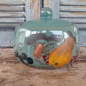 May include: A teal ceramic jar with a lid, hand-painted with a fruit design. The jar features a yellow pear, red accents, and black and blue details. The jar sits on a wooden surface.