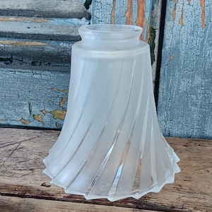 May include: A frosted glass lampshade with a flared, bell-like shape. The shade features a series of vertical, curved lines that create a spiral effect. The glass has a soft, diffused appearance, and the shade is designed to fit over a light bulb.