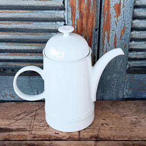 May include: A solid white ceramic coffee pot featuring a cylindrical body, a curved handle, a long tapered spout, and a matching lid with a small knob handle. The base of the pot is decorated with three horizontal embossed rings.