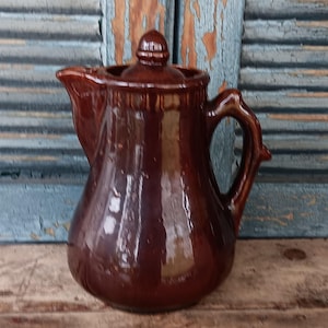 May include: A vintage brown ceramic pitcher with a lid. The pitcher has a curved handle and a spout for pouring. The glossy finish reflects light, and the piece is set against a weathered wooden surface and a blue shutter background.