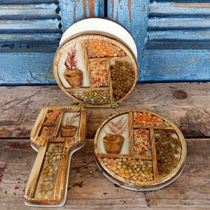 Puede incluir: Un conjunto de tres accesorios de cocina vintage. El conjunto incluye un servilletero redondo, un posavasos y un reposacucharas. Cada pieza presenta un marco de madera con una incrustación de resina transparente llena de varios frijoles secos y una pequeña planta en maceta.