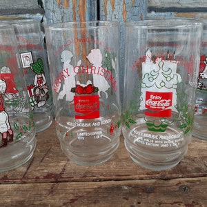 May include: Set of four clear glass Coca-Cola Christmas glasses with red and green illustrations. The glasses feature different holiday scenes, including a Christmas tree, a cow, and children. The glasses are part of the "Holly Hobbie and Robby" series and have the text "Enjoy Coca-Cola" on them.