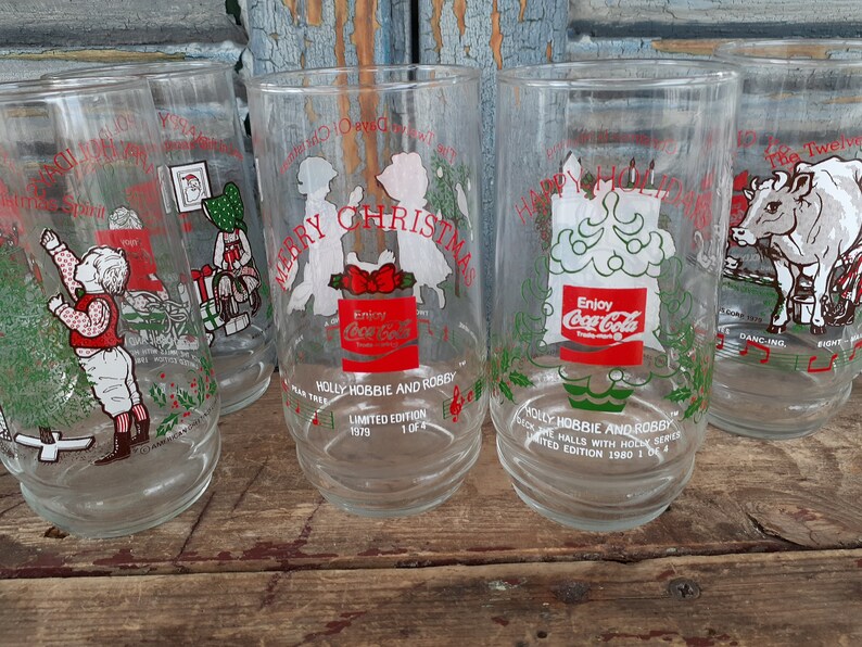 May include: Set of four clear glass Coca-Cola Christmas glasses with red and green illustrations. The glasses feature different holiday scenes, including a Christmas tree, a cow, and children. The glasses are part of the "Holly Hobbie and Robby" series and have the text "Enjoy Coca-Cola" on them.