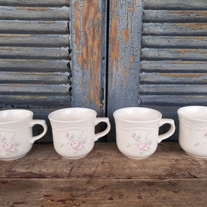 May include: Four white ceramic mugs with a floral design featuring pink roses and green leaves. The mugs have a rounded shape and a handle on the right side.