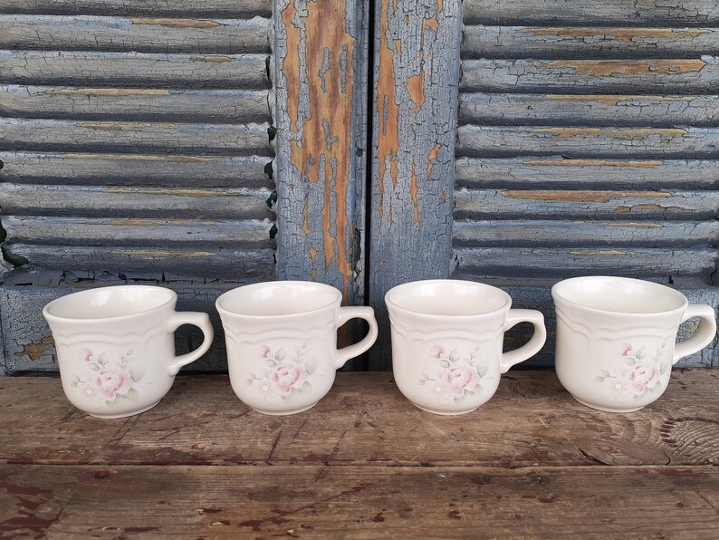 May include: Four white ceramic mugs with a floral design featuring pink roses and green leaves. The mugs have a rounded shape and a handle on the right side.