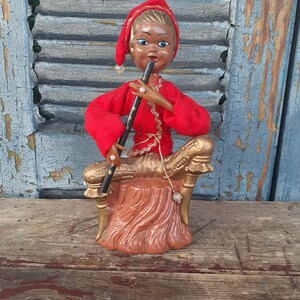 May include: A vintage Christmas figurine of a elf playing a flute, wearing a red and gold outfit, sitting on a brown tree stump.