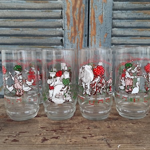 May include: Set of four clear glass drinking glasses with illustrations of the Twelve Days of Christmas. Each glass features a different day of Christmas, with a child and animals. The glasses are on a wooden surface.