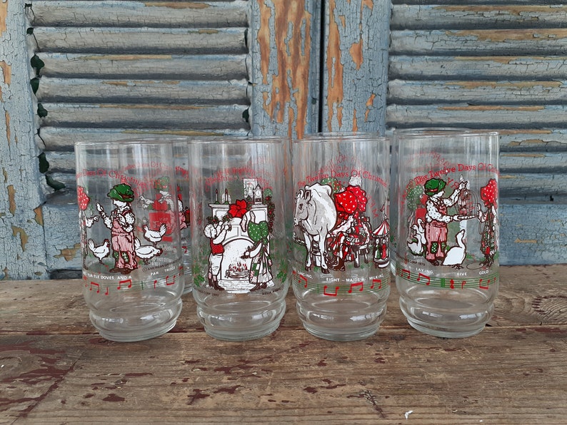 May include: Set of four clear glass drinking glasses with illustrations of the Twelve Days of Christmas. Each glass features a different day of Christmas, with a child and animals. The glasses are on a wooden surface.