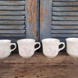 May include: Four white ceramic mugs with a floral design featuring pink roses and green leaves. The mugs have a rounded shape and a handle on the right side.