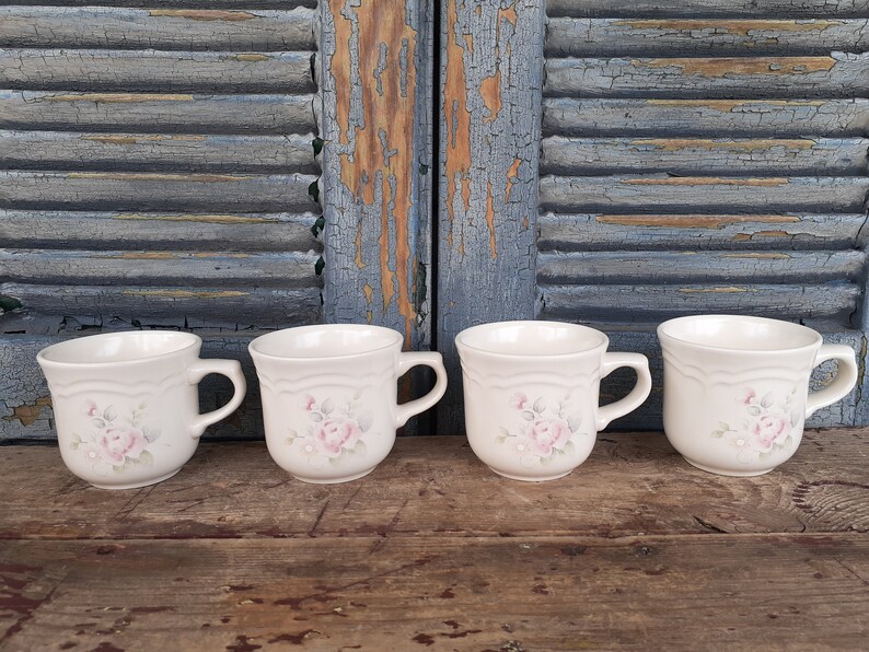May include: Four white ceramic mugs with a floral design featuring pink roses and green leaves. The mugs have a rounded shape and a handle on the right side.