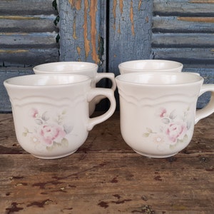 May include: Set of four white ceramic mugs with a floral design featuring pink roses and green leaves. The mugs have a scalloped rim and a handle.