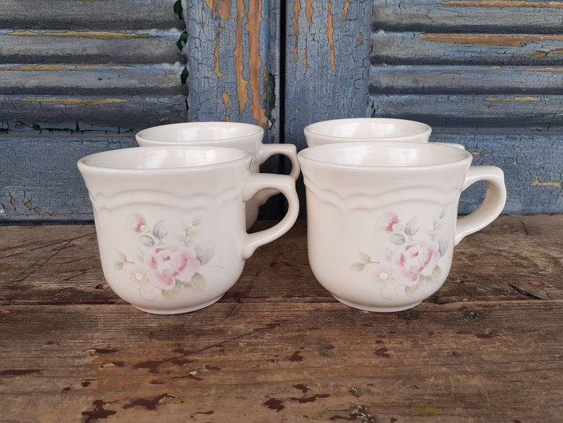 May include: Set of four white ceramic mugs with a floral design featuring pink roses and green leaves. The mugs have a scalloped rim and a handle.